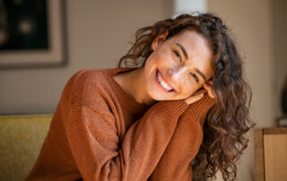 A curly-haired woman wearing a warm brown sweater smiles at the camera.